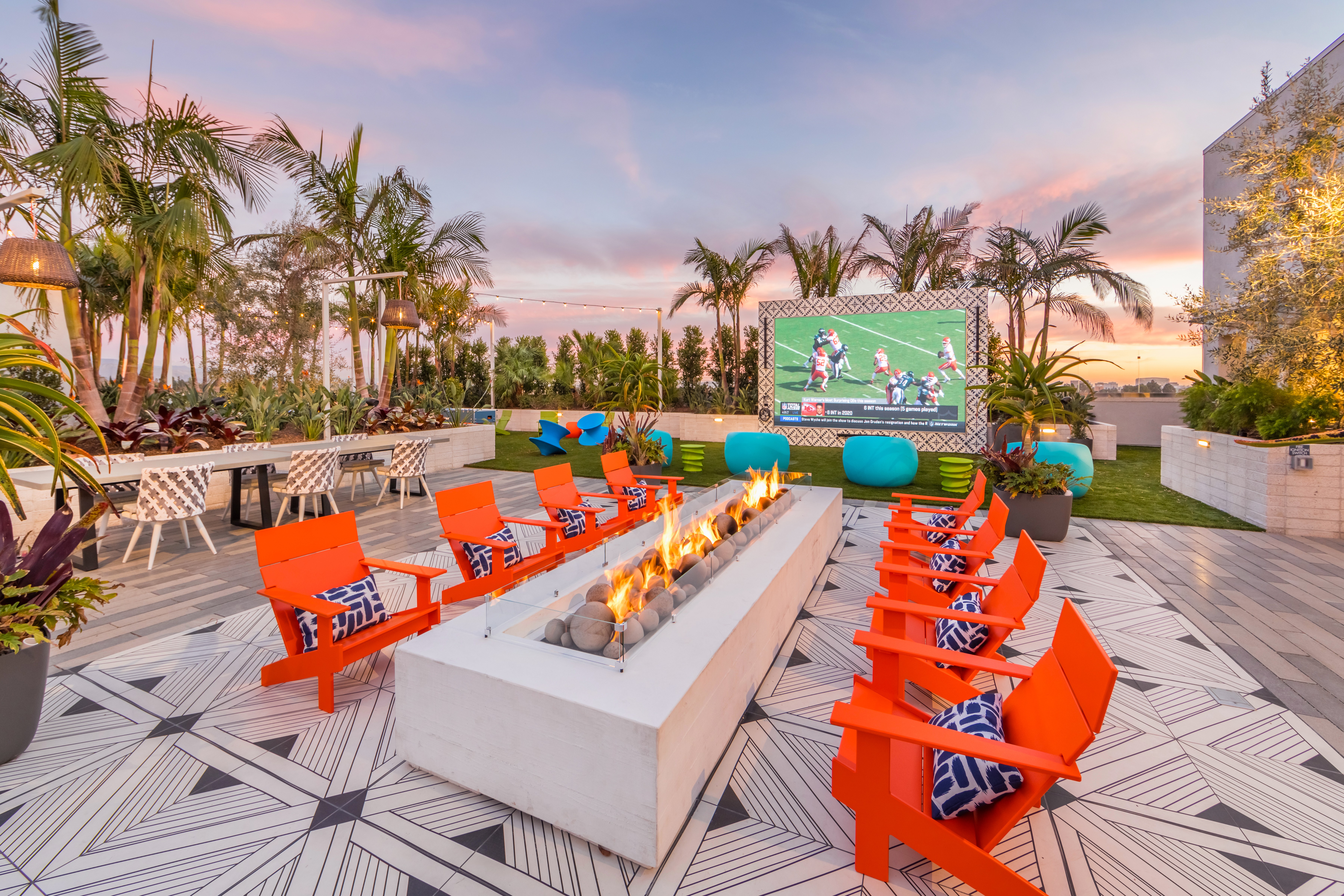 8 Adirondack chairs seated around a long concrete fire table at dusk with palm trees and a large outdoor movie screen in the background