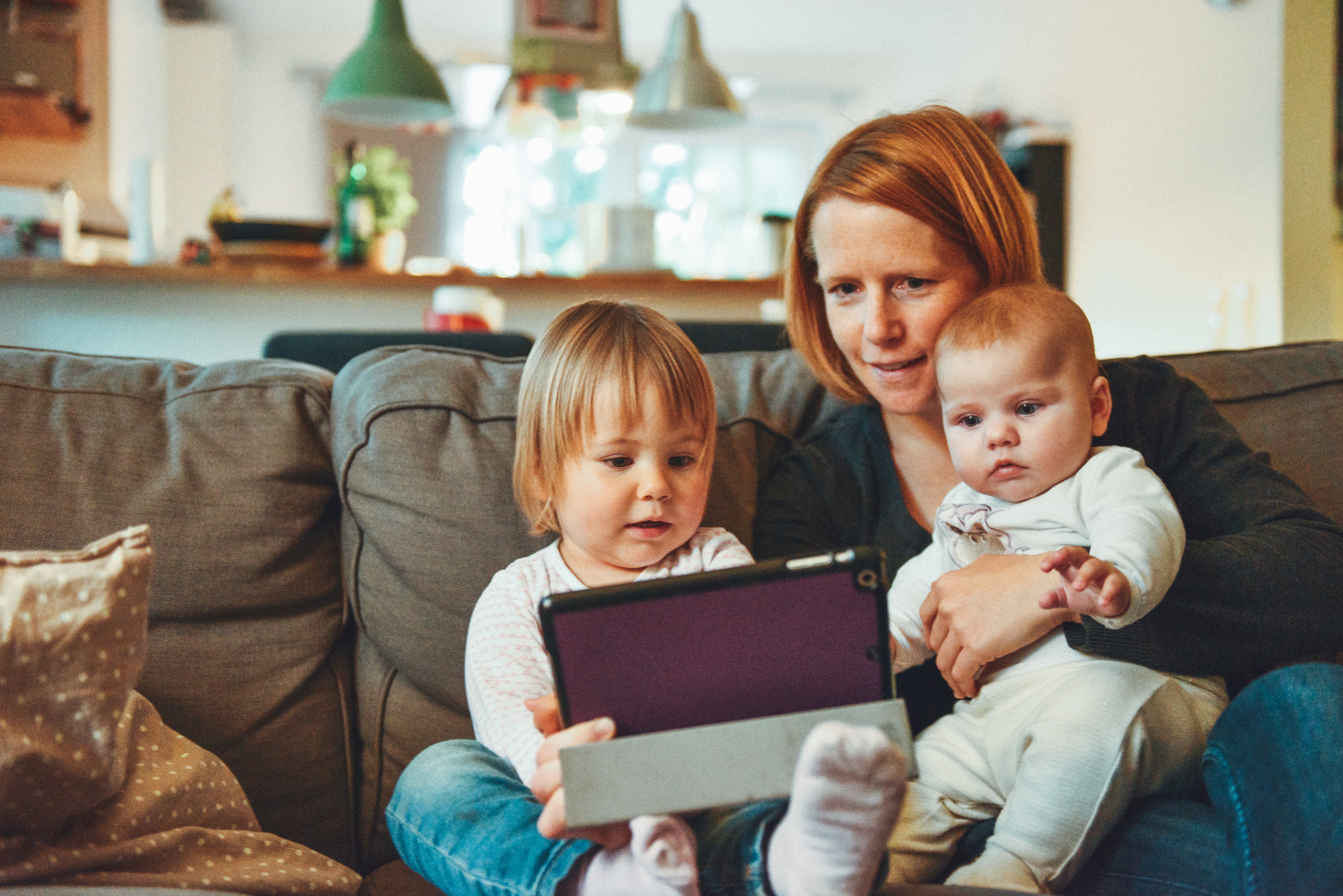 A mother sits on the couch with her toddler and infant.